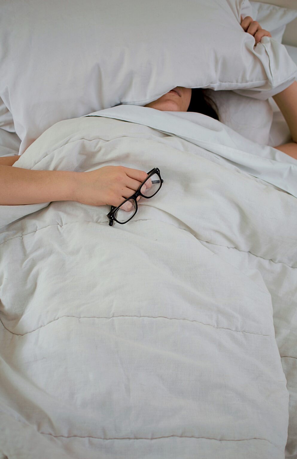 Erschöpfte Person liegt im Bett, hält eine Brille und bedeckt den Kopf mit einem Kissen – Symbol für Burnout und die Notwendigkeit professioneller Hilfe in einer Burnout Klinik.