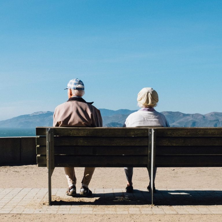 Ein älteres Paar sitzt auf einer Bank mit Blick auf eine weite Landschaft und das Meer. Das Bild symbolisiert Ruhe, Reflexion und die Bedeutung einer ganzheitlichen Senioren Therapie für körperliches und seelisches Wohlbefinden im Alter.